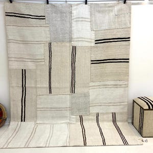 May include: A large, rectangular, patchwork rug with a neutral color palette. The rug features various rectangular patches in shades of beige and gray, with black and gray stripes. A matching ottoman and a round pillow are also visible.