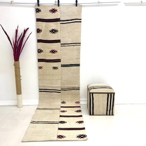 Puede incluir: Alfombra de pasillo beige y burdeos con patrones geométricos y rayas. La alfombra cuelga de un tendedero. A la derecha hay un otomán a juego con rayas negras. A la izquierda hay una planta decorativa con tallos burdeos.
