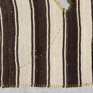 Puede incluir: Un tejido con rayas verticales alternas en marr&oacute;n oscuro y blanco roto. Los bordes y dos recortes irregulares est&aacute;n reforzados con costuras visibles de hilo amarillo en punto de cruz, junto con costuras diagonales amarillas adicionales.