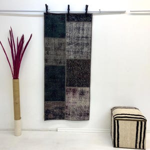 以下が含まれることがあります： グレー、黒、バーガンディの色合いの長方形のパッチワークラグが白い壁に掛けられています。バーガンディの茎が入った背の高い花瓶と、小さなストライプのオットマンも画像にあります。
