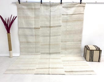 Può includere: Un grande tappeto dai toni neutri con strisce orizzontali in varie tonalità di beige e grigio. Il tappeto è esposto contro un muro bianco, con un vaso decorativo e un pouf a righe nella scena. Il tappeto sembra essere fatto di fibre naturali.