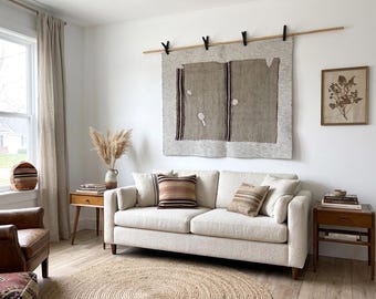 Puede incluir: Un sofá texturizado de color crema con tres cojines decorativos en tonos tierra a rayas, colocado bajo un gran tapiz rectangular tejido suspendido de una barra de madera con cuatro clips negros en una pared blanca lisa.