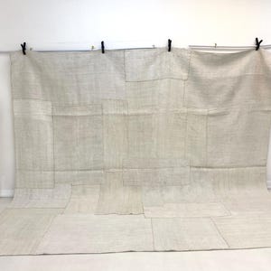 Könnte beinhalten: Ein großer Patchwork-Teppich in neutralen Tönen mit sichtbaren Nähten. Der Teppich besteht aus rechteckigen Stoffstücken in verschiedenen Beige- und Cremetönen. Ein dekoratives Bündel roter Stiele und eine gestreifte Aufbewahrungsbox befinden sich im Hintergrund.