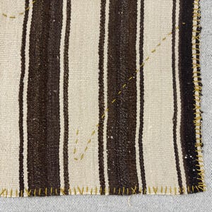 Puede incluir: Un fragmento de tejido con rayas verticales alternas en marr&oacute;n oscuro y blanco roto. Los bordes est&aacute;n rematados con una puntada de sobrehilado visible en color amarillo mostaza, y un hilo amarillo mostaza crea l&iacute;neas decorativas.
