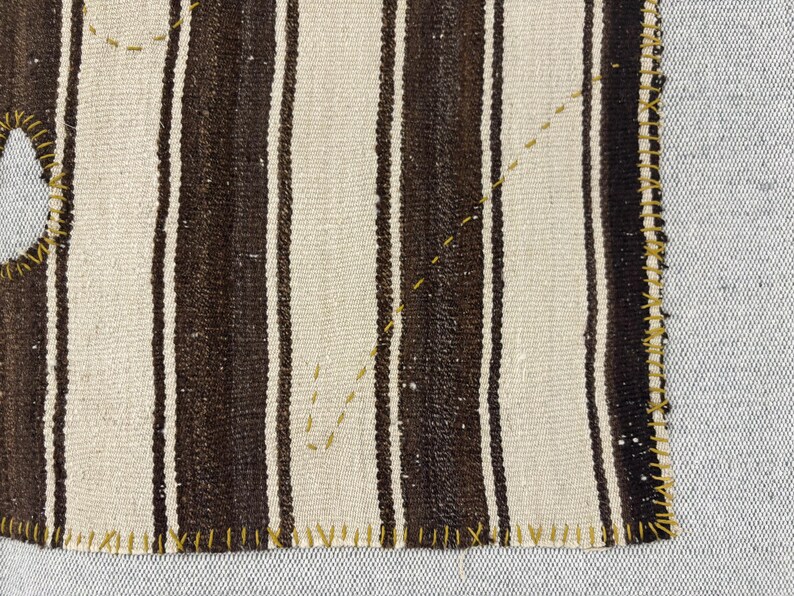 Puede incluir: Un fragmento de tejido con rayas verticales alternas en marr&oacute;n oscuro y blanco roto. Los bordes est&aacute;n rematados con una puntada de sobrehilado visible en color amarillo mostaza, y un hilo amarillo mostaza crea l&iacute;neas decorativas.