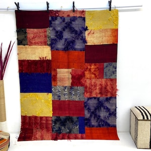 May include: A patchwork textile with a variety of colors, including red, blue, yellow, and gray. The textile is rectangular and appears to be made of different fabric pieces sewn together. It is hanging from a rod with black clips.