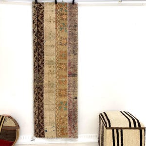 Könnte beinhalten: Ein langer, rechteckiger Teppich mit Patchwork-Design, mit vertikalen Streifen in Braun-, Beige- und Lilatönen. Der Teppich hängt an einer weißen Wand. Ein rundes, gestreiftes Kissen und ein gestreifter Hocker sind ebenfalls sichtbar.