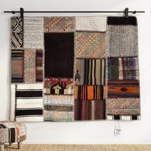 Könnte beinhalten: Ein rechteckiger Wandteppich, der aus verschiedenen zusammengenähten Stoffstücken mit unterschiedlichen Texturen und Farben wie Schwarz, Weiß, Orange und Braun besteht. Das Textil zeigt geometrische Muster, Streifen und einfarbige Blöcke und hängt an einer schwarzen Metallstange.
