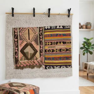 May include: A wall hanging featuring a large, light-colored, textured fabric base suspended from a wooden rod by four black clips. Two distinct, smaller, rectangular woven textile panels with geometric patterns in pink, brown, yellow, and blue are attached to the front of the base fabric.