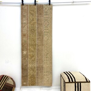 May include: A rectangular, patterned rug in shades of beige, tan, and olive green hangs against a white wall. The rug features a central design with vertical stripes and geometric patterns. A round, patchwork ottoman and a square footstool are also visible.