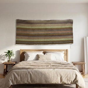 May include: A rectangular wall hanging featuring horizontal stripes in shades of brown, olive green, tan, and muted purple. The textile has a woven texture with visible fringe along the top and bottom edges, displayed horizontally on a plain wall.