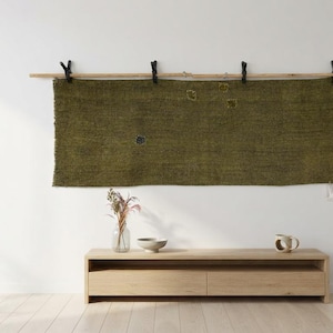 Puede incluir: Un tapiz rectangular de tela tejida en color verde oliva, suspendido de una varilla de madera clara mediante cuatro clips negros. La tela presenta una superficie texturizada con pequeñas manchas dispersas de colores contrastantes y un borde inferior deshilachado.