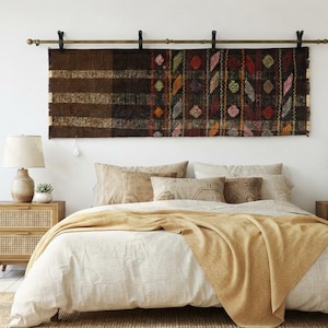 May include: A rectangular woven wall hanging featuring a dark brown section with horizontal stripes and a patterned section with colorful diamond shapes, suspended from a horizontal metal rod with four black mounting brackets above a bed.