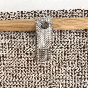 Puede incluir: Un tejido texturizado en tonos marr&oacute;n y blanco roto, con una varilla de madera horizontal insertada a trav&eacute;s de una trabilla de tela. La trabilla est&aacute; asegurada con un ojal met&aacute;lico y cosida al panel textil principal con hilo visible.