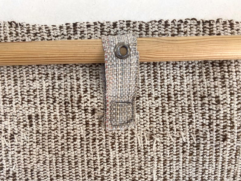 Puede incluir: Un tejido texturizado en tonos marr&oacute;n y blanco roto, con una varilla de madera horizontal insertada a trav&eacute;s de una trabilla de tela. La trabilla est&aacute; asegurada con un ojal met&aacute;lico y cosida al panel textil principal con hilo visible.