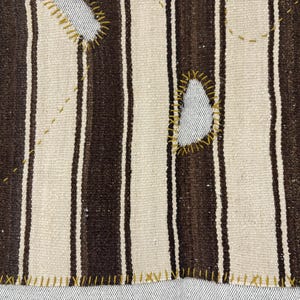 Puede incluir: Un tejido con rayas verticales alternas en marr&oacute;n oscuro y blanco roto. La tela muestra recortes irregulares cosidos a mano y reforzados con hilo amarillo mostaza, junto con puntadas decorativas de color amarillo sobre la superficie.