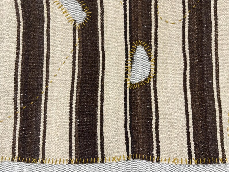 Puede incluir: Un tejido con rayas verticales alternas en marr&oacute;n oscuro y blanco roto. La tela muestra recortes irregulares cosidos a mano y reforzados con hilo amarillo mostaza, junto con puntadas decorativas de color amarillo sobre la superficie.