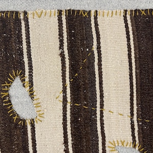 Puede incluir: Un tejido con rayas verticales alternas en marr&oacute;n oscuro y blanco roto. Los bordes y los agujeros irregulares en la tela est&aacute;n reforzados con costuras de hilo amarillo visible, creando un contraste marcado con los tonos neutros.