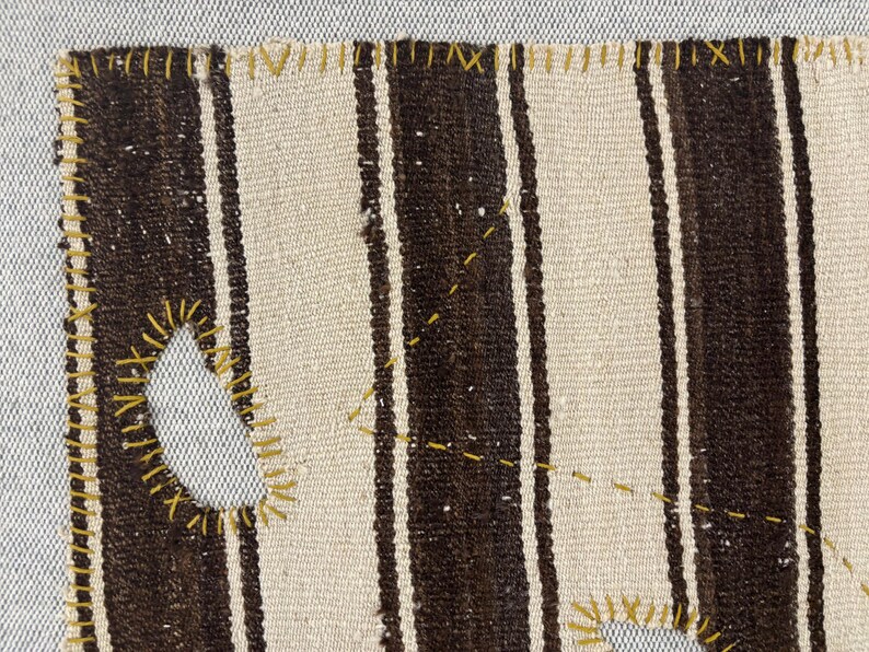 Puede incluir: Un tejido con rayas verticales alternas en marr&oacute;n oscuro y blanco roto. Los bordes y los agujeros irregulares en la tela est&aacute;n reforzados con costuras de hilo amarillo visible, creando un contraste marcado con los tonos neutros.