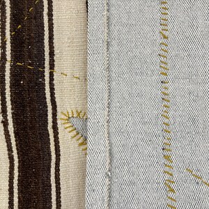 Puede incluir: Una tela textil que presenta dos secciones distintas. El lado izquierdo muestra una base de color crema con gruesas rayas verticales de color marr&oacute;n oscuro y costuras decorativas amarillas. El lado derecho es una tela gris claro lisa con costuras amarillas.