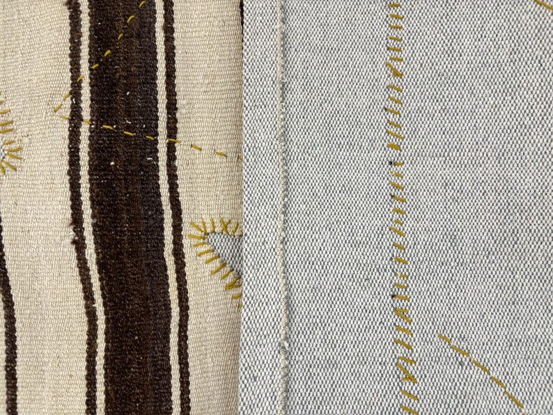 Puede incluir: Una tela textil que presenta dos secciones distintas. El lado izquierdo muestra una base de color crema con gruesas rayas verticales de color marr&oacute;n oscuro y costuras decorativas amarillas. El lado derecho es una tela gris claro lisa con costuras amarillas.