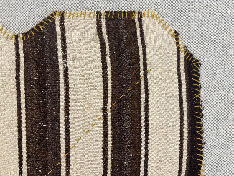Puede incluir: Una pieza de tela tejida con rayas verticales alternas en marr&oacute;n oscuro y blanco roto. Los bordes est&aacute;n rematados con una puntada de sobrehilado amarilla visible, y una l&iacute;nea diagonal de costura decorativa amarilla cruza el centro del tejido.