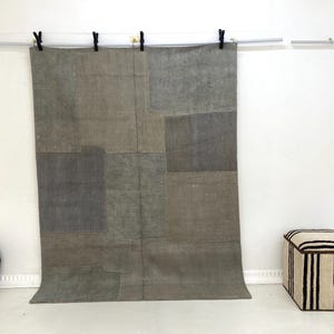 May include: A large, rectangular, patchwork rug in shades of gray and beige hangs from a metal rod with black clips. A striped ottoman and a round, patterned cushion sit nearby against a white wall. The rug's design features various rectangular patches.