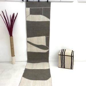 Puede incluir: Una alfombra rectangular larga con un diseño geométrico en tonos grises y blanquecinos cuelga de un tendedero. La alfombra presenta una serie de rayas horizontales y formas abstractas. Un jarrón decorativo con plumas burdeos está a la izquierda.