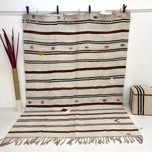 May include: Cream-colored, striped rug with dark red, black, and gold horizontal bands. The rug has a fringed edge and small geometric patterns. It is hanging from a metal rod with black clips.