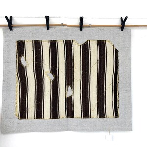 Puede incluir: Una pieza rectangular de tela gris claro con un textil rectangular m&aacute;s peque&ntilde;o centrado, con rayas verticales de color marr&oacute;n oscuro y crema. La secci&oacute;n rayada tiene tres agujeros visibles y est&aacute; cosida a la base gris con hilo amarillo.