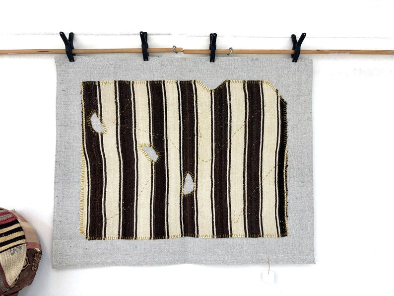 Puede incluir: Una pieza rectangular de tela gris claro con un textil rectangular m&aacute;s peque&ntilde;o centrado, con rayas verticales de color marr&oacute;n oscuro y crema. La secci&oacute;n rayada tiene tres agujeros visibles y est&aacute; cosida a la base gris con hilo amarillo.