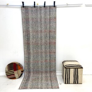 Puede incluir: Una alfombra larga y rectangular gris con rayas rojas cuelga de una barra. Un cojín redondo de retazos y una otomana a rayas están en el suelo. La alfombra está hecha de un material tejido.