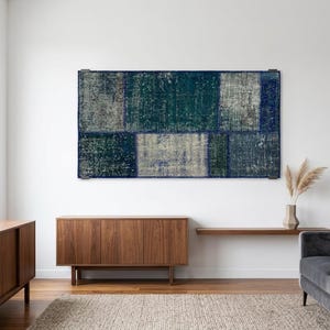 Puede incluir: Un tapiz de pared rectangular con un diseño de patchwork de paneles de tela desgastados en tonos azul marino profundo, verde azulado y gris apagado. La obra de arte textil está montada horizontalmente en una pared blanca, mostrando costuras visibles entre las variadas secciones rectangulares que crean una apariencia texturizada y multitonal en toda la superficie.