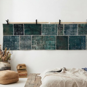 May include: A long, rectangular wall hanging composed of multiple patchwork squares in varying shades of deep teal, forest green, and dark blue. The textile features a distressed, weathered texture and is suspended from a wooden rod with black clips.