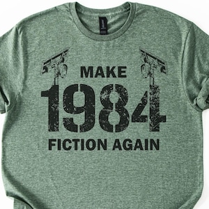 May include: A heathered sage green short-sleeve t-shirt featuring a black graphic print. The design includes two security cameras flanking the text "MAKE 1984 FICTION AGAIN" in a distressed, bold, sans-serif font centered on the chest area.