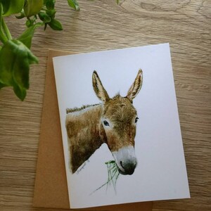 May include: A watercolour painting of a brown donkey with large ears, eating green grass. The donkey is looking directly at the viewer.