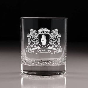May include: A clear, engraved rocks glass featuring a family crest design. The crest includes two lions, a hand symbol, and the name "Devaney" on a banner. The glass has a textured base and is suitable for whiskey or other beverages.
