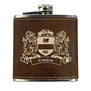May include: A brown leather-wrapped flask with a silver cap and trim. The flask features a family crest design with two lions, a shield, and the name "Conlon" in a banner. The flask is rectangular and has a hinged cap.