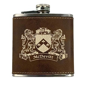 May include: A brown leather flask with a silver cap and trim. The flask features a detailed crest with two lions, a shield, and the name "McDevitt" on a banner. The design is in a lighter color, contrasting with the dark brown leather.