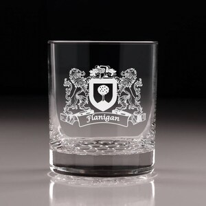 May include: A clear, etched glass tumbler featuring a family crest design. The crest includes two lions, a shield with a tree, and a banner with the name "Flanigan". The glass has a textured base and is suitable for beverages.