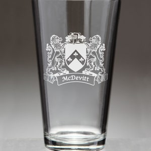 May include: A clear pint glass with a white family crest design. The crest features a shield with three clover-like symbols, flanked by two lions. The name "McDevitt" is displayed on a banner below the crest.