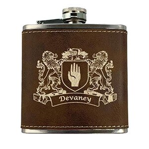 May include: A brown leather-wrapped flask with a silver cap and trim. The flask features a family crest design with two lions, a hand, and the name "Devaney" engraved on the front.