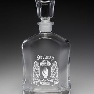 May include: A clear glass decanter with a square base and a stopper. The decanter features the name "Devaney" and a family crest with a hand symbol and the word "Ireland" etched in white.