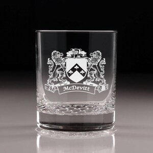 May include: A clear, engraved rocks glass featuring a family crest design. The crest includes a shield with three clover shapes, flanked by two lions, and the name "McDevitt" on a banner. The glass has a textured base.