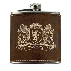 May include: A brown leather flask with a silver cap and trim. The flask features a family crest design with two lions, a shield, and the name "McCrary" engraved on the front.