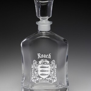 May include: A clear glass decanter with a square body and a stopper. The decanter features the name "Roach" above a family crest with three fish and the word "Ireland" below.
