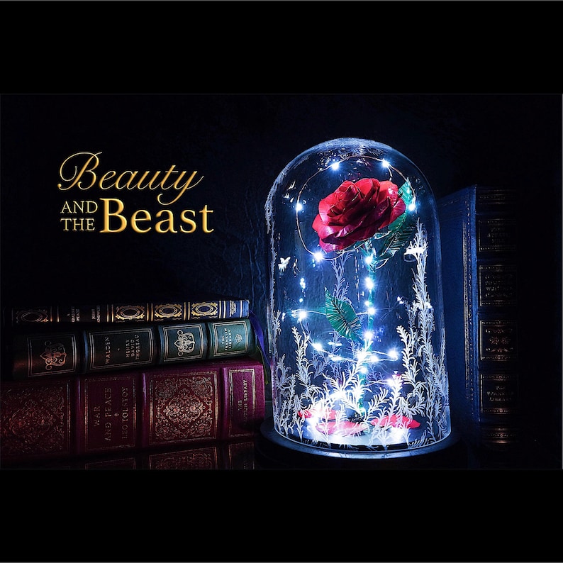 Op de afbeelding: Een decoratieve glazen stolp met een rode roos en fee&euml;rieke lichtjes. De basis is zwart en de stolp is versierd met een bloemenpatroon. De tekst "Beauty and the Beast" staat in goud. Boeken op de achtergrond.