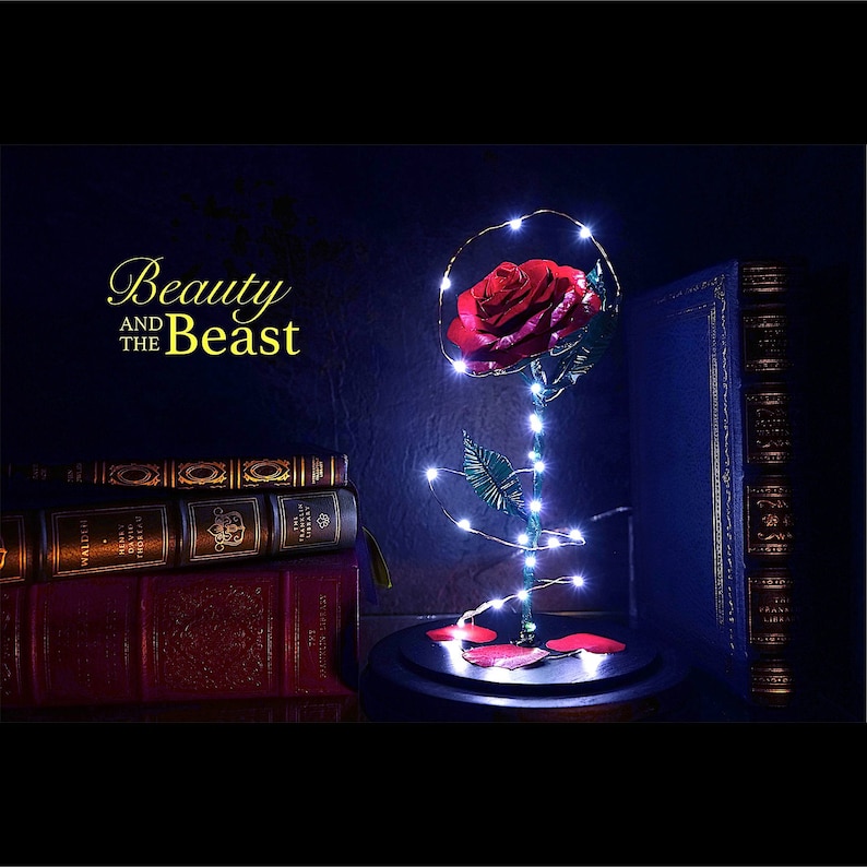 Op de afbeelding: Een rode roos in een glazen stolp, verlicht door fee&euml;rieke lichtjes, met de gouden tekst "Beauty and the Beast". De roos staat op een zwarte voet, met boeken op de achtergrond.