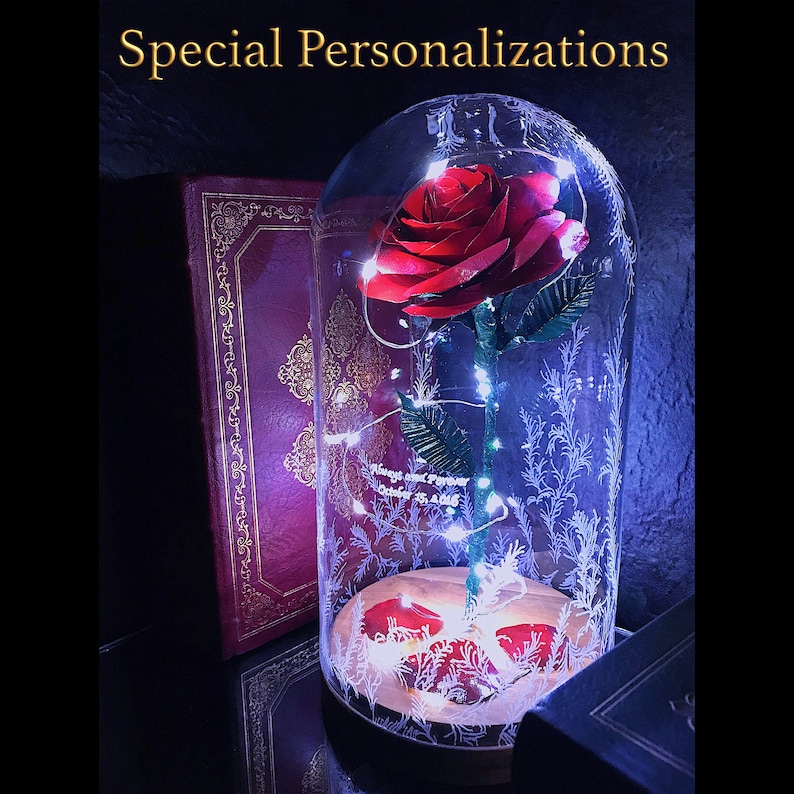 Op de afbeelding: Een rode roos in een glazen stolp met fee&euml;rieke lichtjes, met de tekst "Special Personalizations". Een rood boek is zichtbaar op de achtergrond. De glazen stolp heeft een gegraveerd ontwerp en de woorden "Always and Forever October 25, 2016".