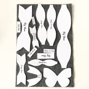 May include: A set of white felt patterns for making bows and butterflies. The patterns include different sizes for big and small bows, as well as a butterfly pattern. The patterns are labeled with the names of the different bow sizes and the words "Bow w Tails".